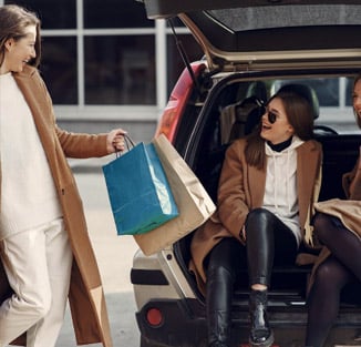 a woman holding shopping bags is talking to a woman seated in the open boot of her car